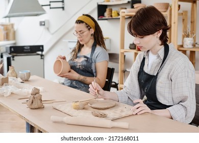 Young Woman In Workwear Sitting By Table And Cutting Ornament On Clay Workpiece Against Colleague With Bowl Creating New Products