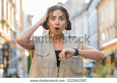 Young woman worrying to be punctual, with anxiety checking time on watch, running late to work or transport, being in delay, deadline outdoors. Girl tourist walking in city street. Town lifestyles