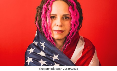 Young woman wrapped in American flag on red background. Female looking at camera, posing in studio. Concept of Flag Day, national holiday