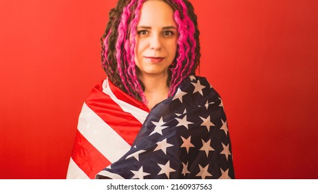 Young woman wrapped in American flag on red background. Female looking at camera, posing in studio. Concept of Flag Day, national holiday