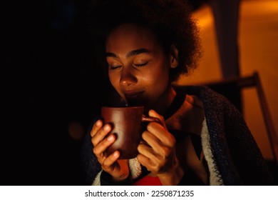 Young woman wrapped in blanket drinking tea while sitting by bright tent in forest at night - Powered by Shutterstock