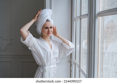 Young Woman Wrapped In Towel And White Robe Standing Next To Bedroom Window Drying Her Hair After A Morning Shower. Smiling Lady With Towel On Her Head Looking At Empty Space, Home Interior, Panorama