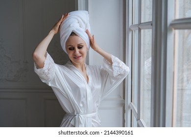 Young Woman Wrapped In Towel And White Robe Standing Next To Bedroom Window Drying Her Hair After A Morning Shower. Smiling Lady With Towel On Her Head Looking At Empty Space, Home Interior, Panorama