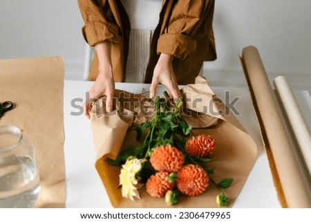 A young woman wraps a bouquet of orange dahlias in kraft paper for a gift. Flower shop, small business and flower delivery concept.