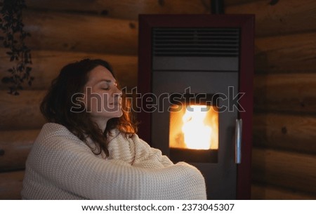 A young woman wraps herself in a warm blanket while sitting by a pellet stove in a wooden cabin. Warm home in cold winter concept