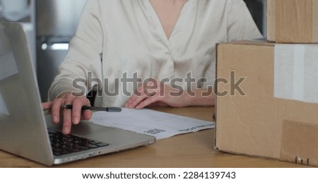 A young woman writes a return order and sits at a table with a laptop at home. Close-up video of a female customer writing on paper and filling out a form, wanting to return cardboard box back.