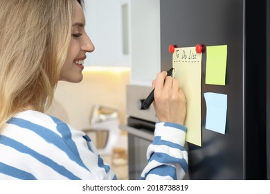 Young Woman Writing To Do List On Refrigerator Door In Kitchen