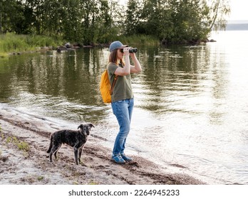 Young woman with yellow backpack looking through binoculars at birds on lake and walking mixed breed dog bedlington terrier whippet Birdwatching, zoology, ecology Ornithology wildlife manager