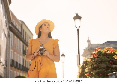 Young woman in a yellow dress and sunhat holding a vintage camera while standing outdoors. She looks up with a curious expression, enjoying a sunny day.  - Powered by Shutterstock