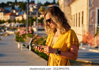 Young woman in yellow dress is using her smartphone while walking down sunny city street in coastal town. Female tourist checks her smartphone while standing by marina at sunset - Powered by Shutterstock