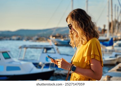 Young woman in yellow dress is using her smartphone while walking down sunny city street in coastal town. Female tourist checks her smartphone while standing by marina at sunset - Powered by Shutterstock