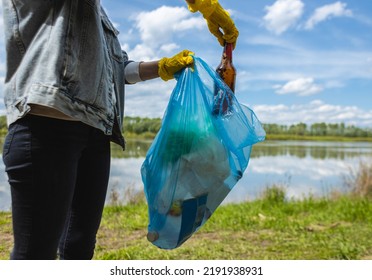 A young woman in yellow gloves collects abandoned garbage in a black bag in the forest. Plastic pollution and environmental protection - Powered by Shutterstock