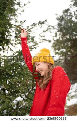 A young woman in a yellow hat is reaching for a Christmas tree branch and having fun on a snowy background. Winter activities and holidays concept. Vertical image