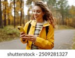 Young woman in a yellow jacket checks her phone while walking on a forest path during autumn