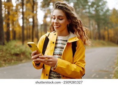 Young woman in a yellow jacket checks her phone while walking on a forest path during autumn