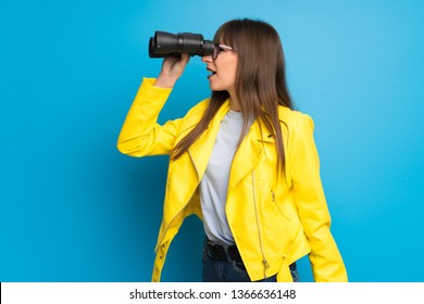 Young woman with yellow jacket on blue background and looking in the distance with binoculars