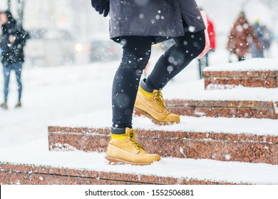 A Young Woman In Yellow Leather Boots Rises On Icy Snowy Granite Steps In Urban Environment. Winter Walk. Abstract Winter Weather Background