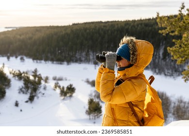 Young woman in yellow looking through binoculars at birds on snowy river against winter forest Birdwatching, zoology, ecology Research in nature, observation of animals Ornithology copy space