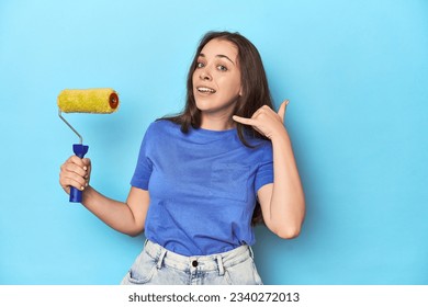 Young woman with yellow paint roller on a blue background showing a mobile phone call gesture with fingers. - Powered by Shutterstock