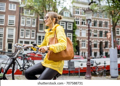 Young woman in yellow raincoat with bag and flowers riding a bicycle in Amsterdam city