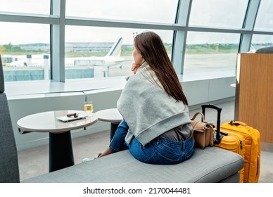 Young woman with yellow suitcase sitting in airport business lounge waiting for plane departure enjoying glass of white vine looking at window at plane.