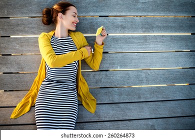 Young woman in yellow sweater using yellow phone on wooden sunbed, top view with space for your text