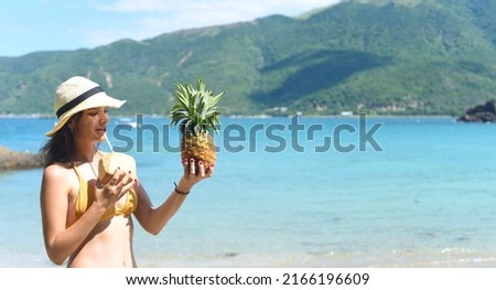 Young woman in yellow swimsuit drinking coconut milk and holding pineapple on sandy beach