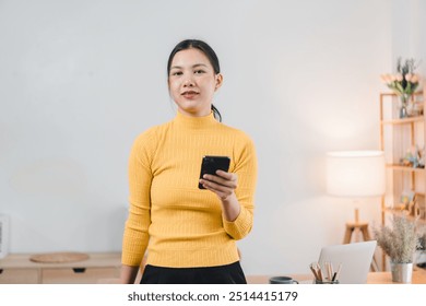 A young woman in yellow turtleneck sweater stands confidently holding smartphone in modern workspace, exuding sense of professionalism and focus. - Powered by Shutterstock