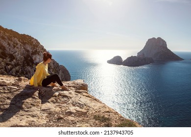 young woman in yellow zip jacket sitting on a cliff looking out on the Mediterranean Sea from Ibiza to famous Es Vedra. High quality photo