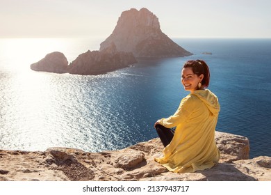 young woman in yellow zip jacket sitting on a cliff turning back to the camera with famous view from Ibiza to Es Vedra in the background. High quality photo