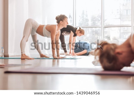 Young woman yoga instructor doing asana shows the correctness of doing to her students in the gym with a large window. Energy channel opening and therapeutic effect concept