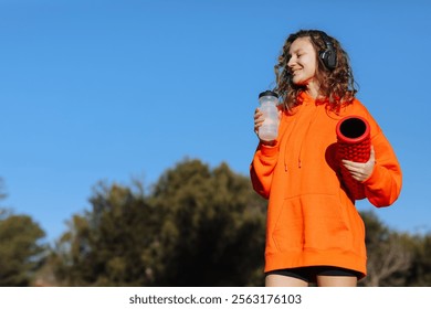 Young woman in with yoga roller drinks water from bottle, listens music in headphones, rests after workout outdoor. Fitness, sport, healthy active lifestyle. Break, hydration, wellness, aqua balance - Powered by Shutterstock