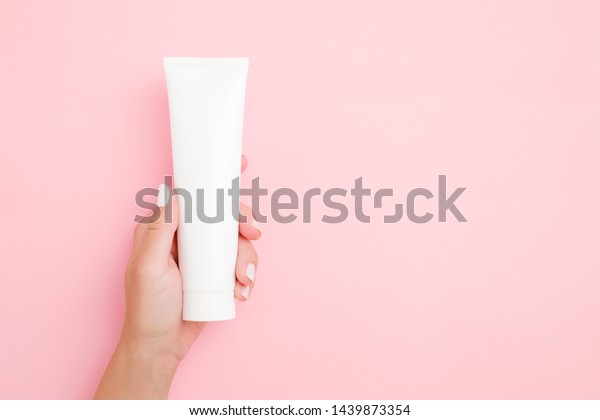 Young\
woman\'s hand holding and showing white tube on light pink table.\
Pastel color. Care about clean and soft body skin. Daily beauty\
product. Close up. Empty place for text or\
logo.