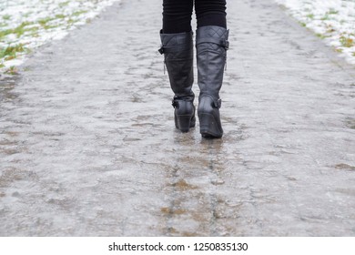 Young Woman's Legs In Black Leather Boots Walking On Sidewalk In Wet, Warm Winter Day. Pavement Covered With Slippery Ice. Back View. Closeup. 