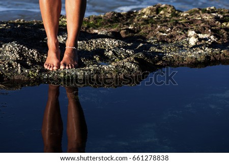 A young woman's toned legs and bare feet are reflected in a calm rock pool as she explores an Australian coastline at low tide.