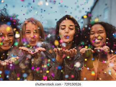 Young women blowing confetti from hands. Friends celebrating outdoors in evening at a terrace.