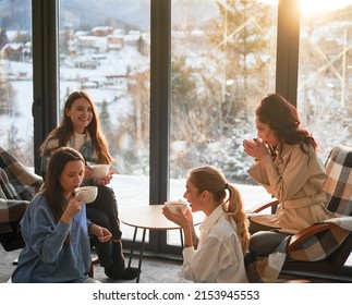 Young Women Enjoying Winter Weekends Inside Contemporary Barn House. Four Girls Having Fun And Drinking Hot Tea Near Panoramic Windows At Sunset.