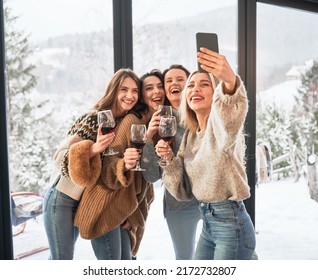 Young Women Enjoying Winter Weekends Inside Contemporary Barn House. Four Girls Having Fun, Taking Selfies On Phone And Drinking Wine.