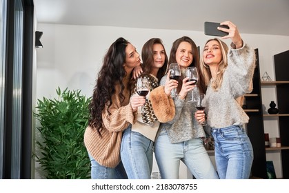 Young Women Enjoying Winter Weekends Inside Contemporary Barn House. Four Girls Having Fun, Taking Selfies On Phone And Drinking Wine.