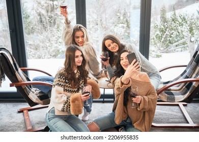 Young Women Enjoying Winter Weekends Inside Contemporary Barn House. Four Girls Having Fun, Taking Selfies On Phone And Drinking Wine.