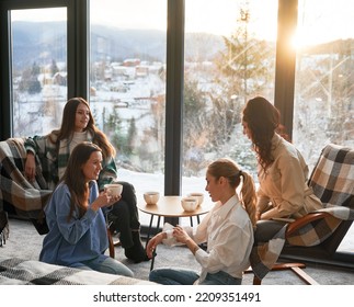 Young Women Enjoying Winter Weekends Inside Contemporary Barn House. Four Girls Having Fun And Drinking Hot Tea Near Panoramic Windows At Sunset.