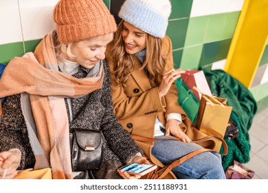 Young women friends shopping together in winter, using smartphone while surrounded by colorful shopping bags. Image showcases fashion, technology use, and friendship in a vibrant urban setting. - Powered by Shutterstock