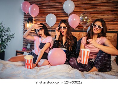 Young Women Have Discgussed Emotions On Face. They Watch Movie Through Special Glasses. Models Sit On Bed In Festive Room. They Have Pajamas Party.