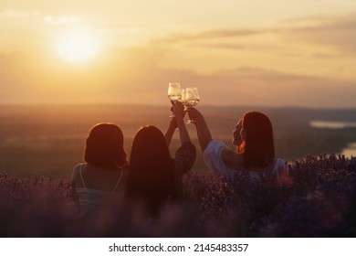 Young Women Having Nice Picnic In The Lavender Field, Cheerful Talk, Clink Glasses With Wine On Bright Summer Sunset. Cheers. Concept Of Party, Joy, Fun And Friendship.