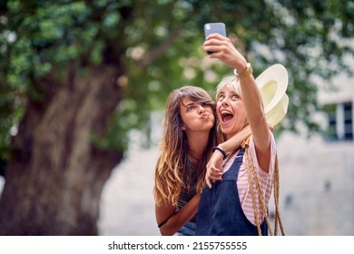 Young Women Making Funny And Goofy Face Expressions And Taking Selfie, On Street. Young Women In European City. Travel, Fun, Togetherness Concept.