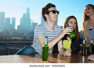 Young Women And Man Drinking Beer And Laughing At Rooftop Bar With Los Angeles Skyline, USA