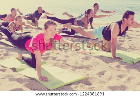 Young women and men exercising yoga poses on sunny beach by ocean