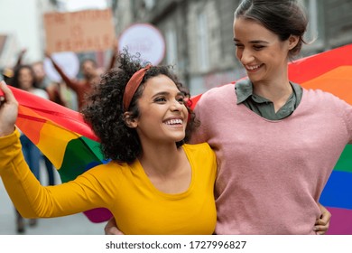 Young women on street enjoying holding gay pride flag during protest. Smiling multiethnic women enjoying during march on street for lgbt rights. Diversity, tolerance and gender identity concept.