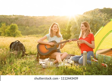 Young Women Resting With Hot Drink And Guitar Near Camping Tent In Wilderness