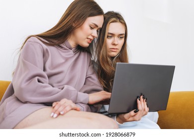 Young Women Sisters Twins Teenagers In Hoodie Beautiful Girls Using Laptop Sitting On Yellow Couch In The Modern Interior